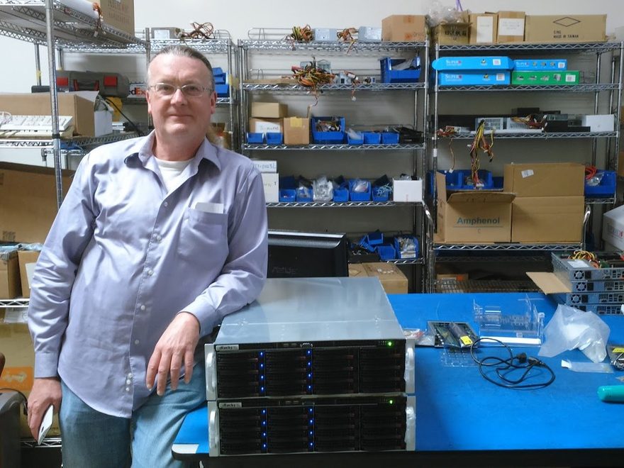 Joe Wolff standing at a workbench beside two rackmount storage servers, in a workshop lined with parts shelving.