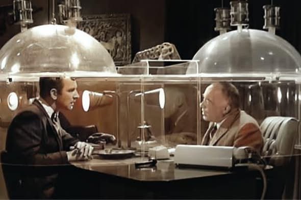 Two men seated at a table, each under a clear plastic dome lowered from the ceiling, leaning in to hear each other.