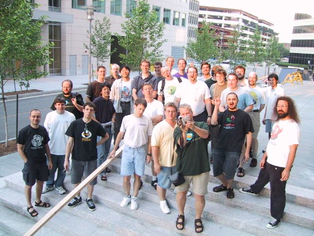 About thirty OpenBSD developers posed together on the outdoor steps of a building, in daylight.